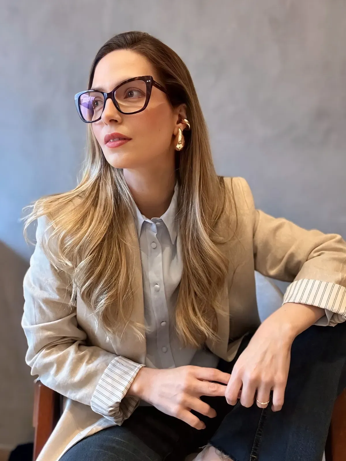 Dra. Amanda Arnoni, de óculos e blazer bege, sentada e olhando para o lado