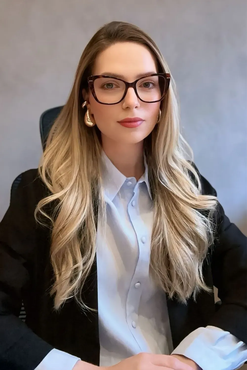 Retrato da Dra. Amanda Arnoni, de óculos, camisa azul-clara e blazer escuro, olhando para a câmera